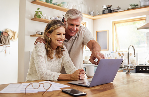 A photo of a retired couple in their kitchen looking at their investments on their laptop.